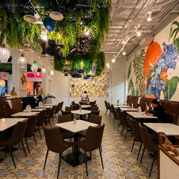 Bright restaurant dining room with white tables, brown booths, hanging greenery, colorful mural, patterned tile floor, and diners.