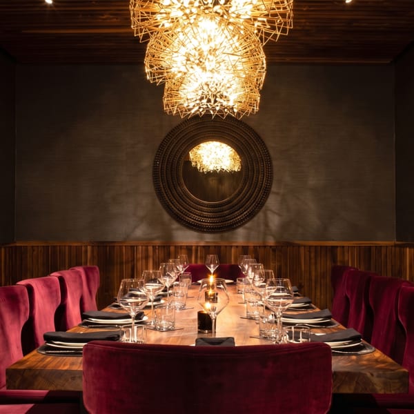 Elegant restaurant dining room with a candlelit wooden table, burgundy velvet chairs, colorful wall art, and sculptural chandeliers.