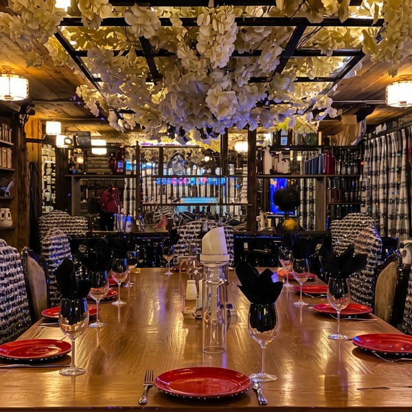 Warmly lit restaurant dining room with a long set table, red plates, wine glasses, patterned chairs, and floral ceiling decorations.
