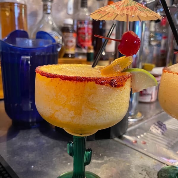 Citrus cocktail with a chili-salt rim, lime wedge, orange slice, cherry, and paper umbrella in a cactus-shaped glass.