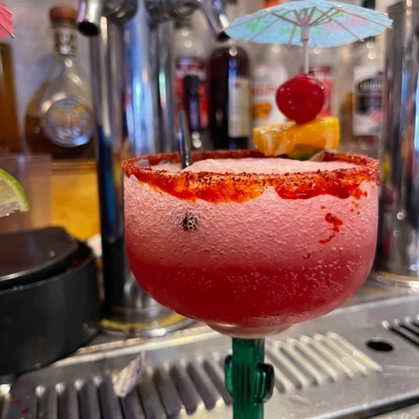 Red cocktail with a chili-salt rim, citrus garnish, and decorative umbrella served at a busy restaurant bar.