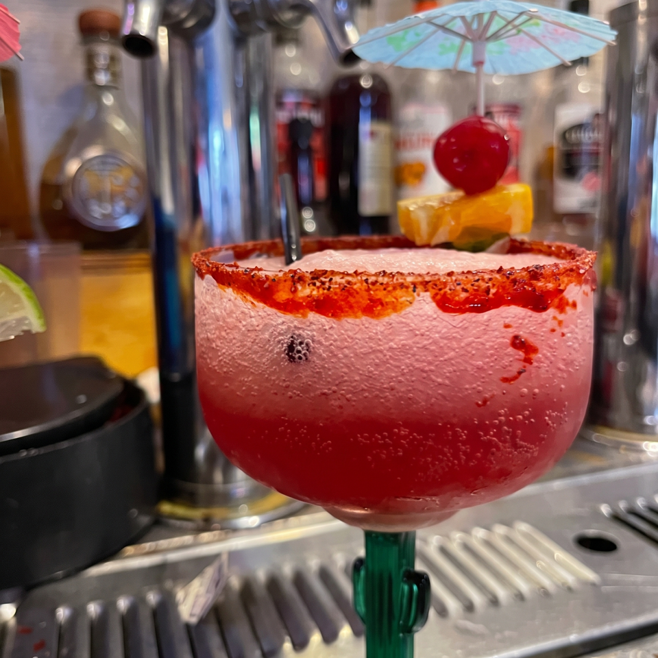 Red cocktail with a chili-salt rim, citrus garnish, and decorative umbrella served at a busy restaurant bar.